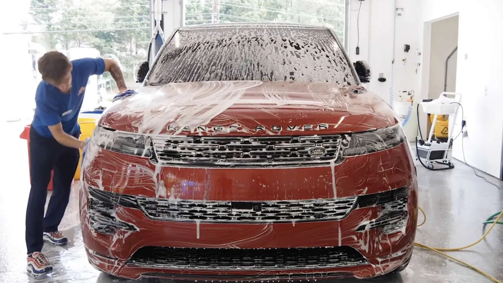 Person washing a red Range Rover SUV covered in soap foam inside a bright garage.