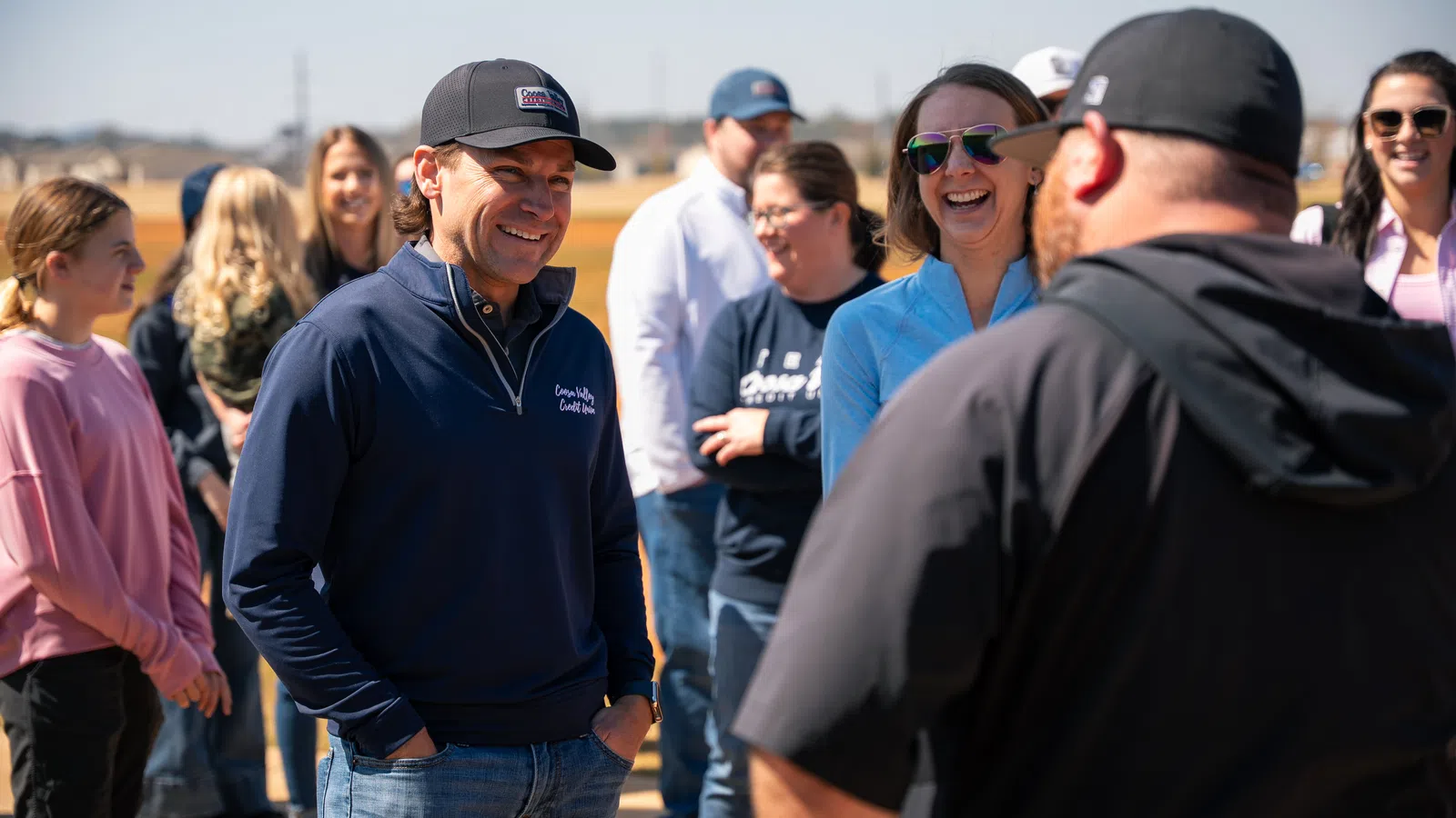 Group of people smiling and chatting outdoors on a sunny day with blue skies and casual attire