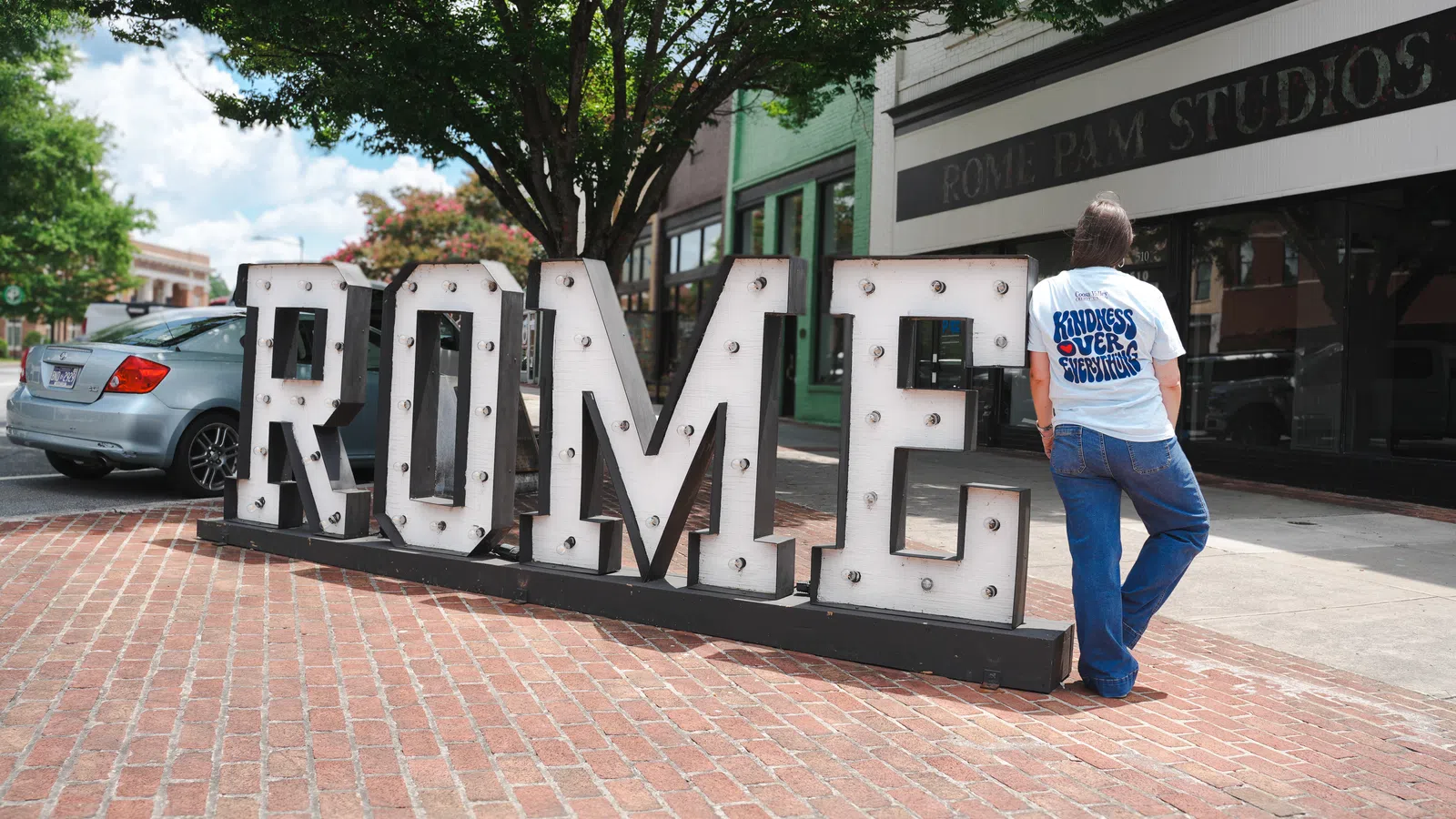Person leaning on large illuminated letters spelling ROME on a brick sidewalk outside Rome PAM Studios.