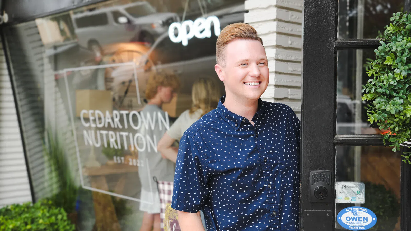 Smiling man stands at the entrance of Cedartown Nutrition store with open sign and greenery outside