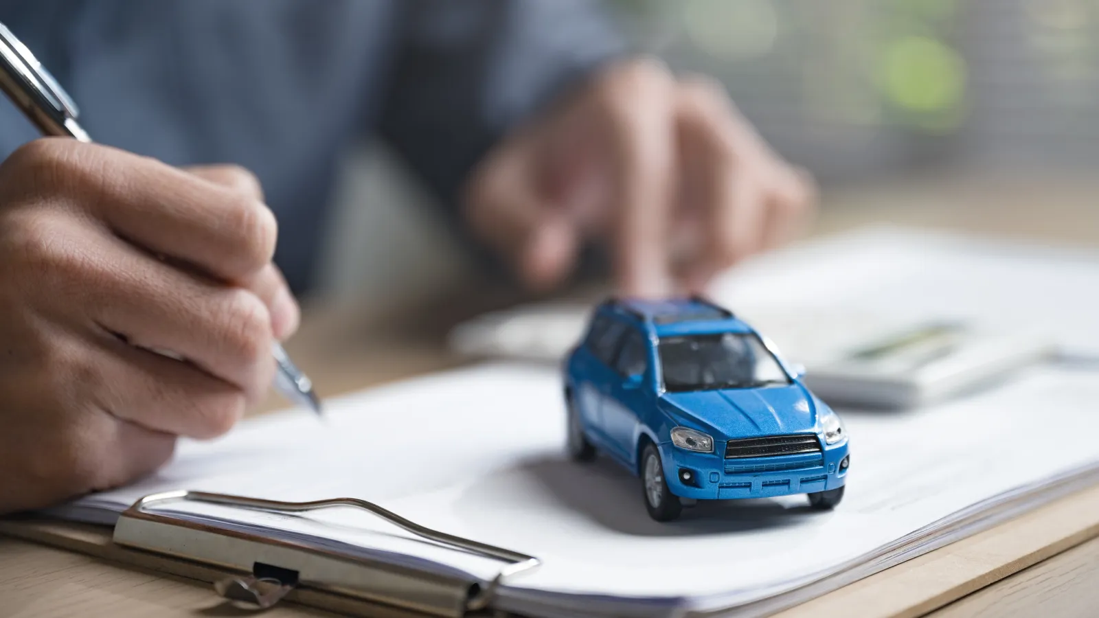 Person signing documents on clipboard with blue toy car symbolizing car insurance or purchase.