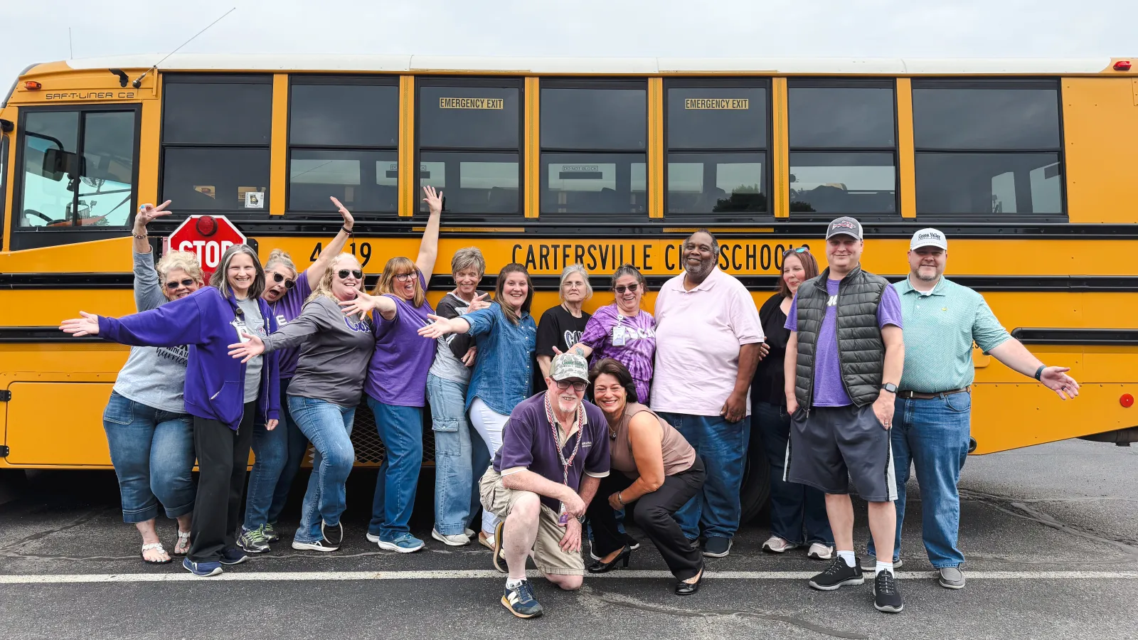 Group of cheerful adults posing in front of a yellow Cartersville City School bus in a parking lot.