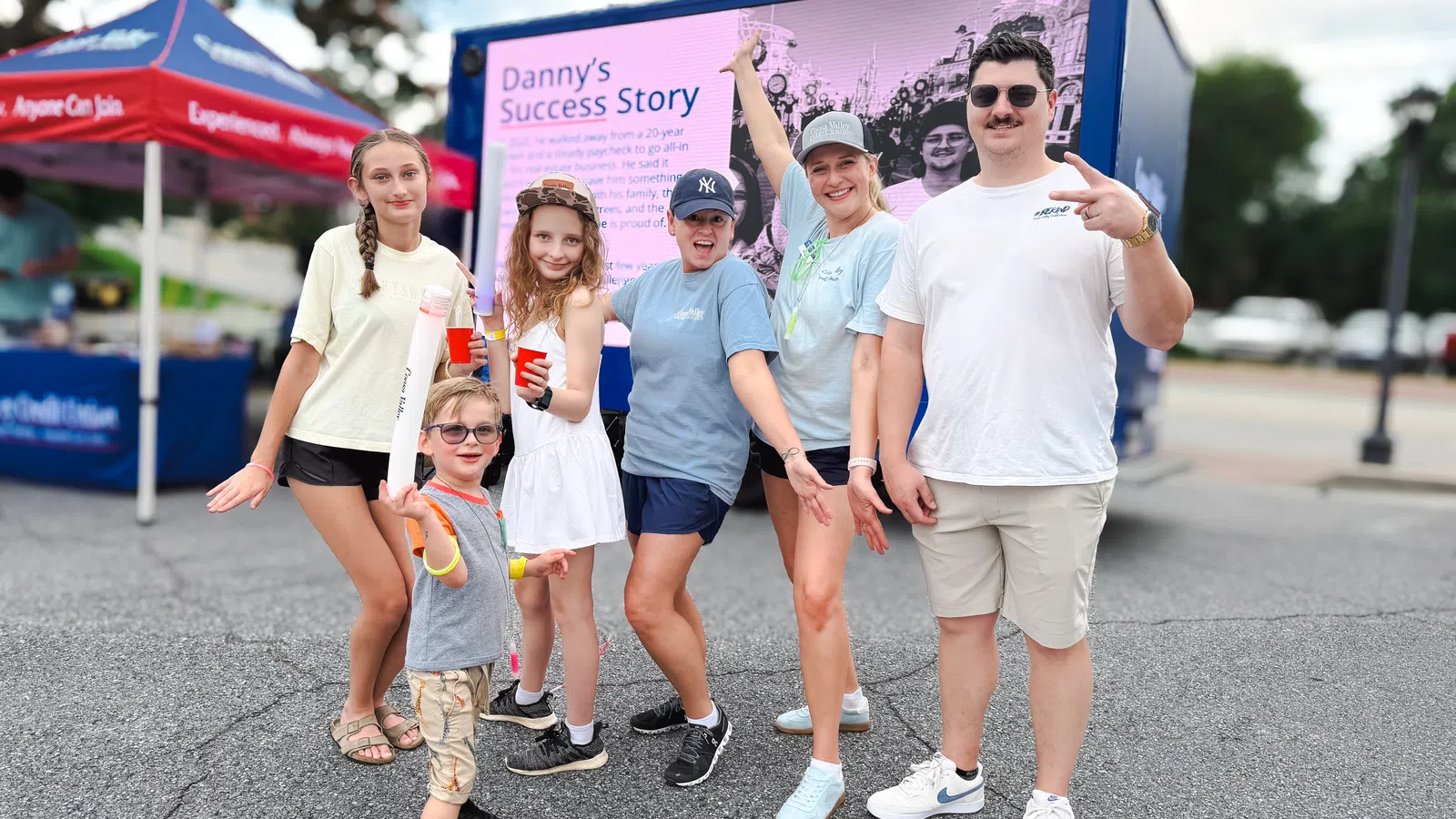 Group of smiling people posing outdoors near a colorful event display with casual summer clothing and lively expressions.