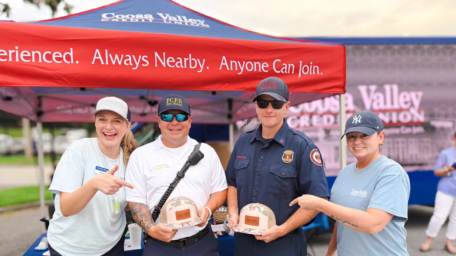 Four people, including two firefighters holding helmets, smiling under a red Coosa Valley Credit Union tent at an outdoor event.