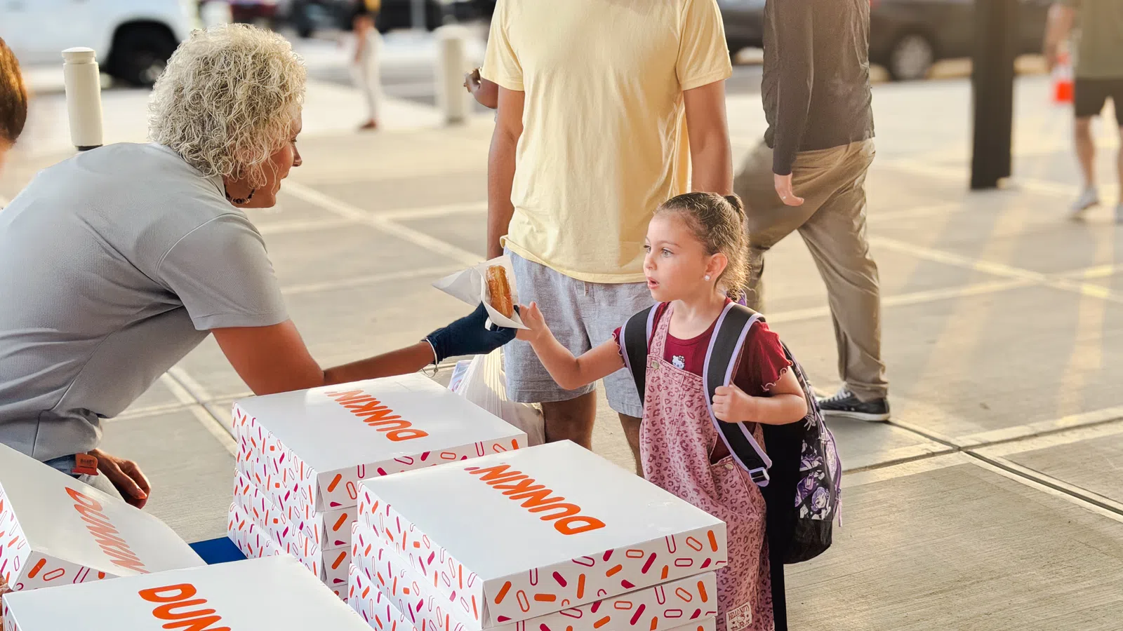 Young girl with backpack receives a Dunkin' donut from a vendor at an outdoor event with her father nearby.