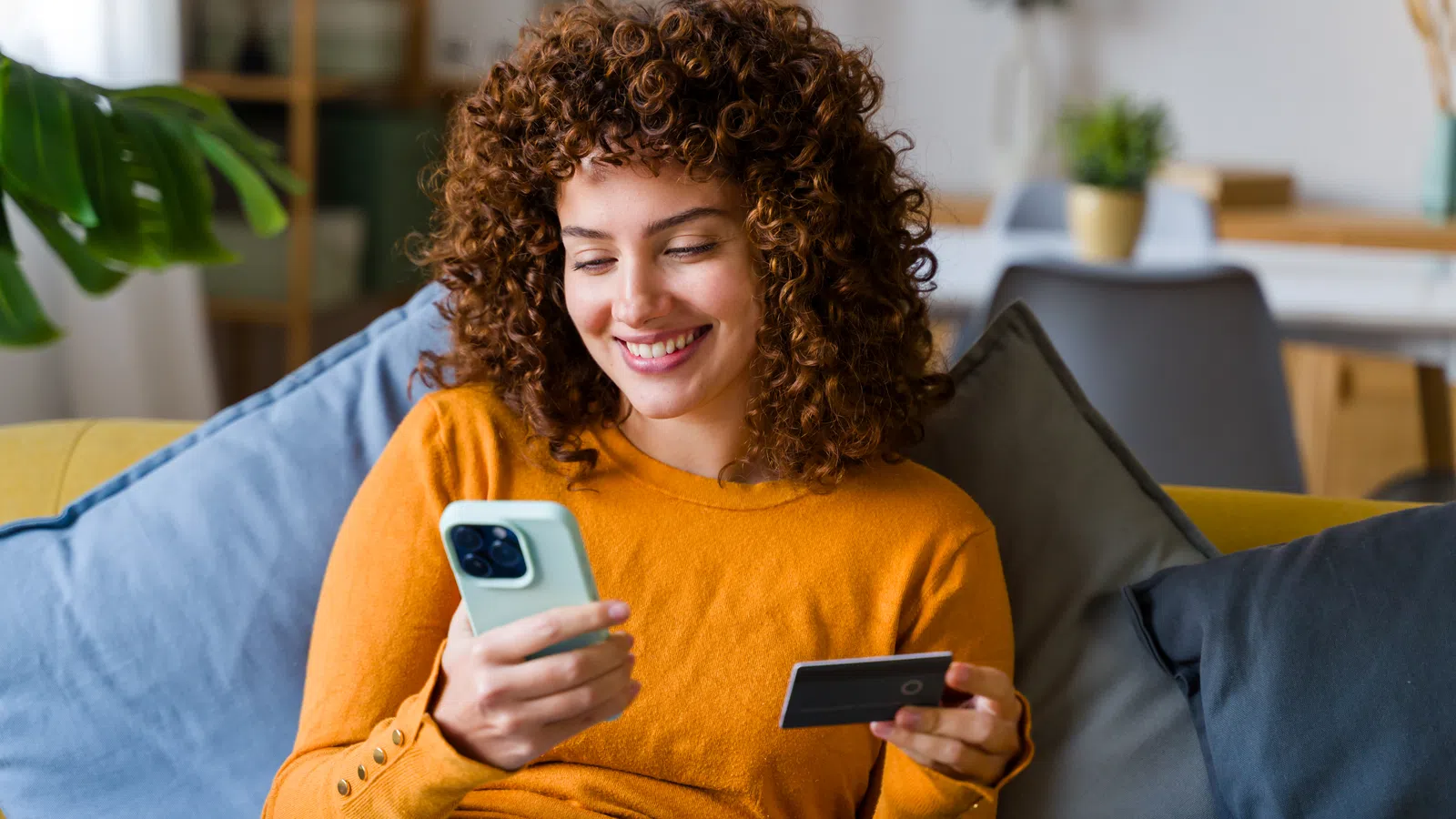 Smiling woman with curly hair using smartphone and credit card for online shopping at home.