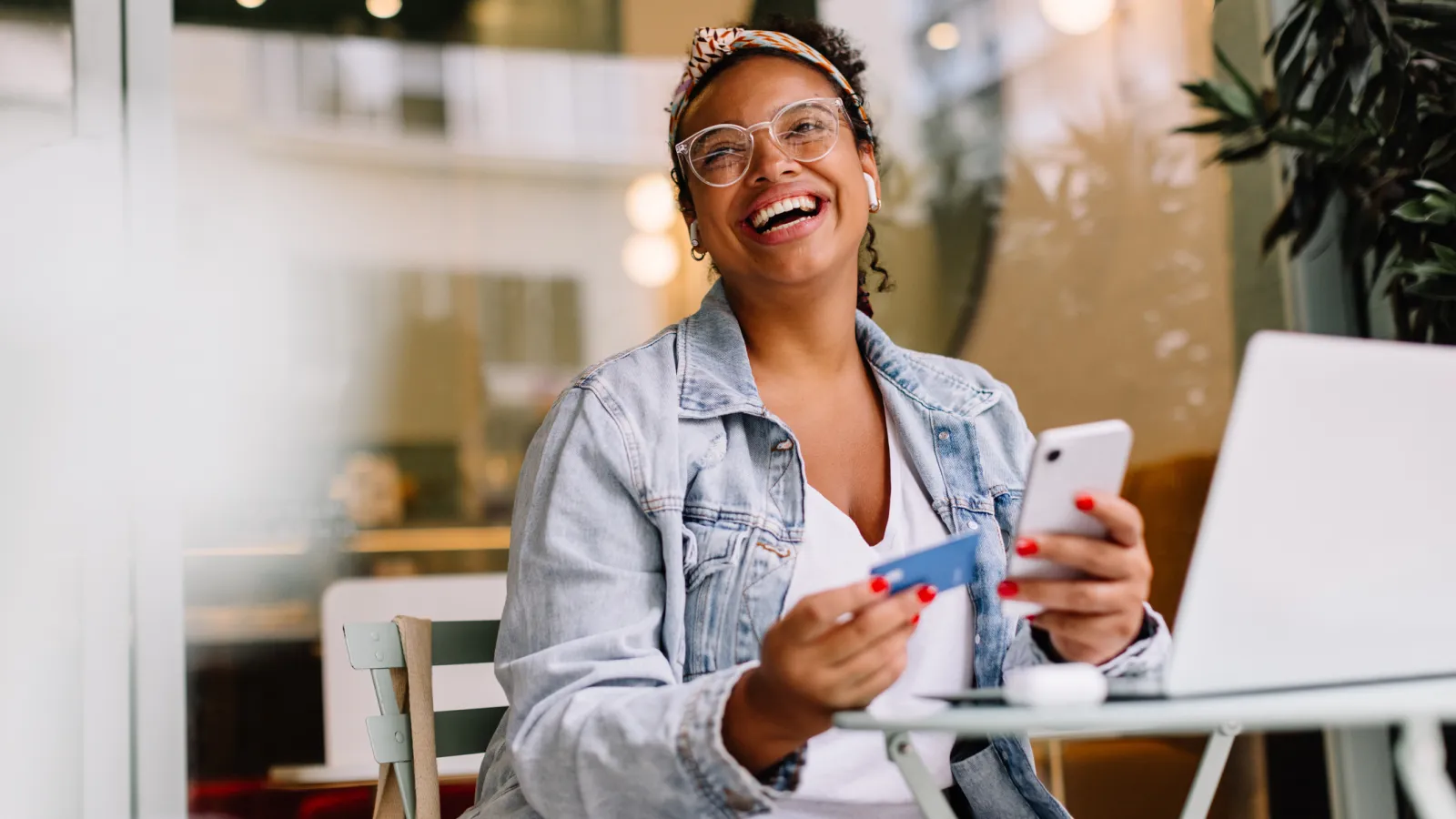 Smiling woman in glasses and denim jacket holds credit card and smartphone near laptop in bright cafe.
