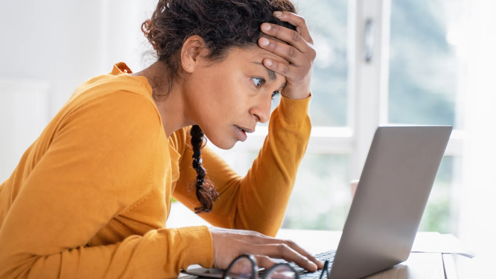 Stressed woman in an orange sweater looking at a laptop screen with worry at a desk near a window.