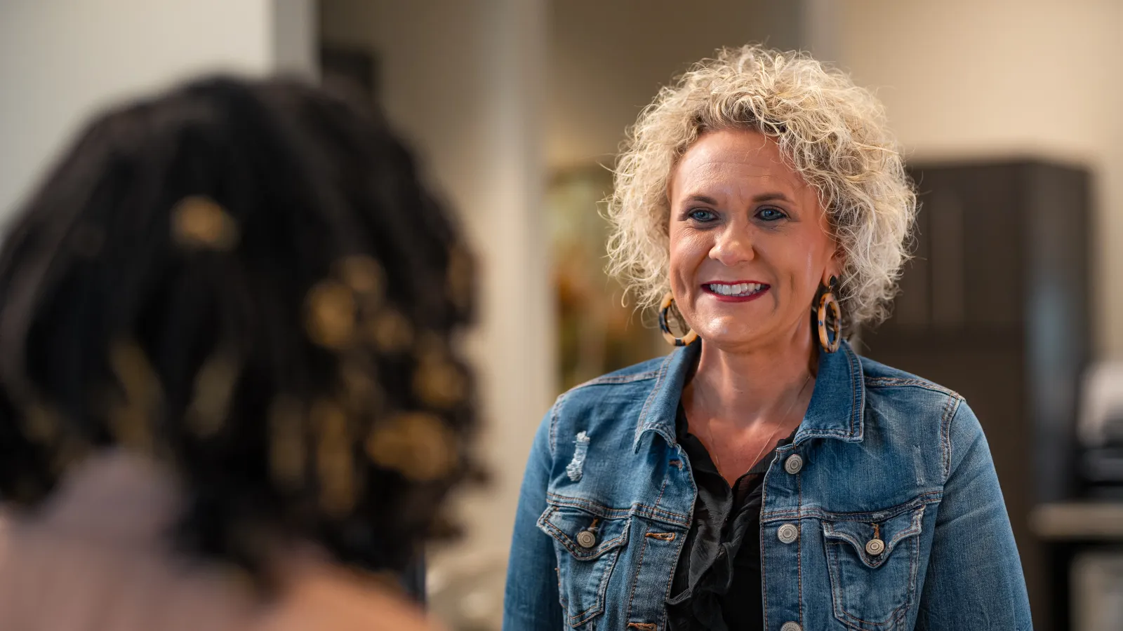 Smiling woman in a denim jacket engaging in conversation with another person indoors.