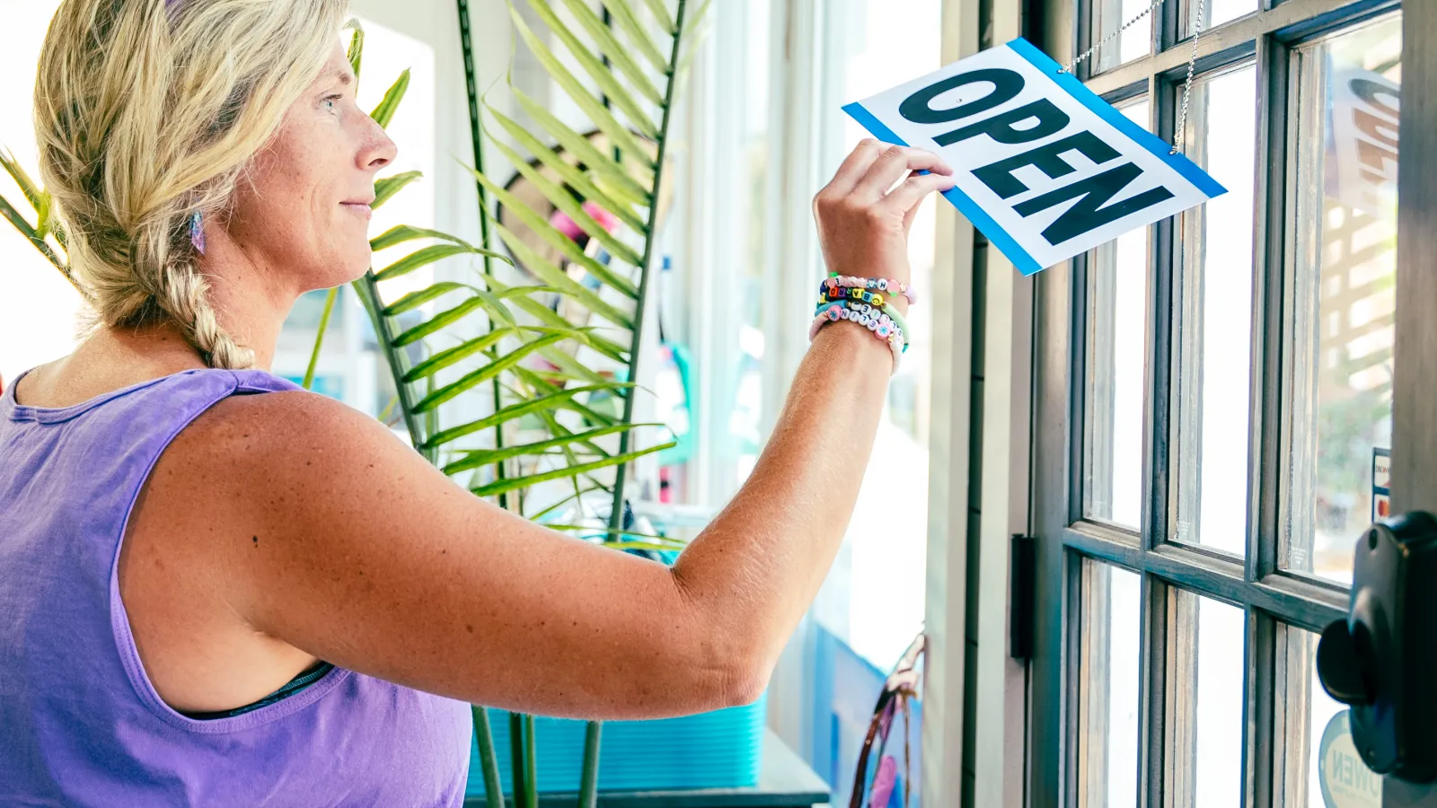 Woman hanging an open sign on a glass door preparing to open a small business in bright daylight.