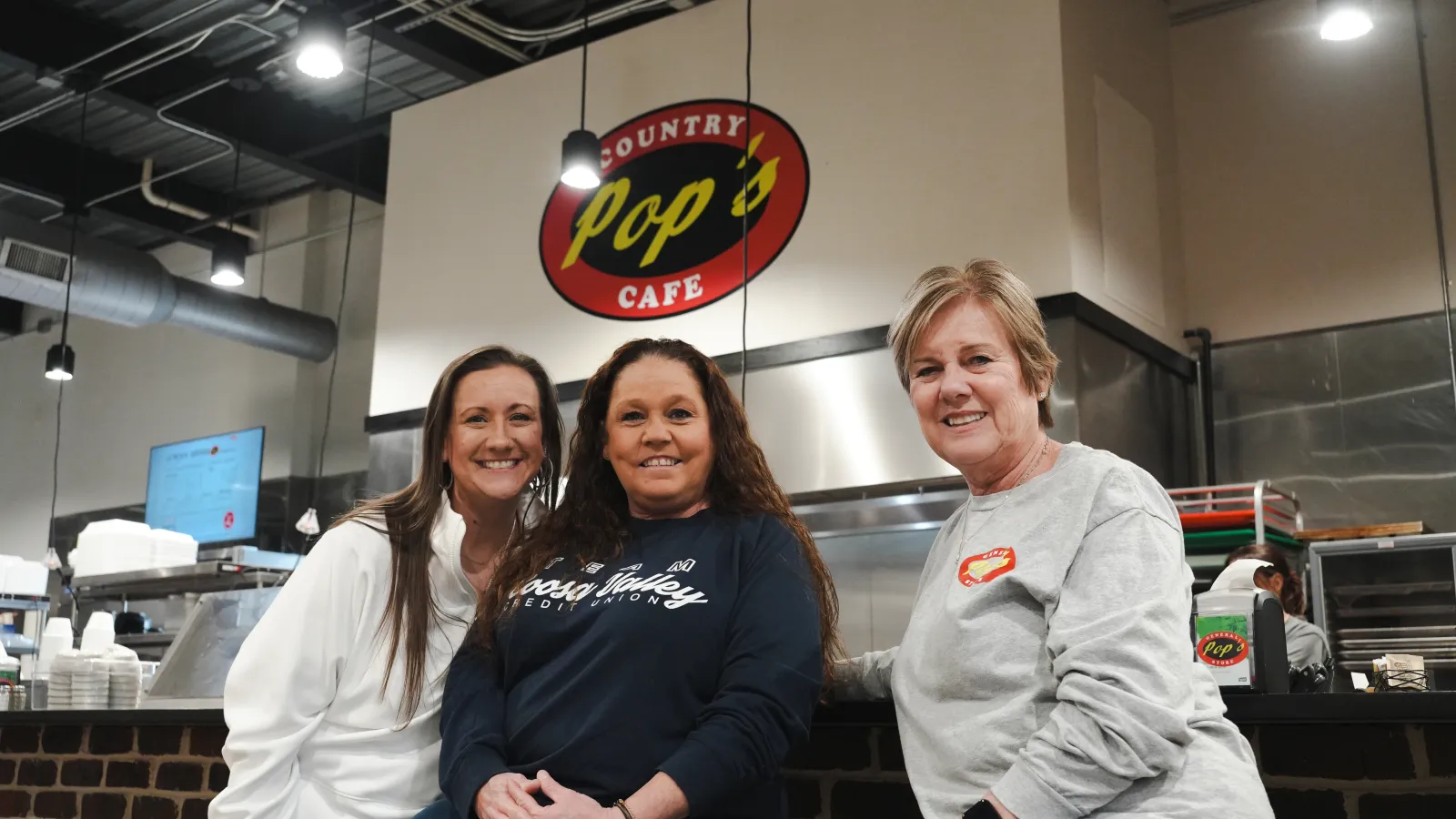 Three women posing inside Pop's Country Cafe with visible kitchen and cafe sign in background.