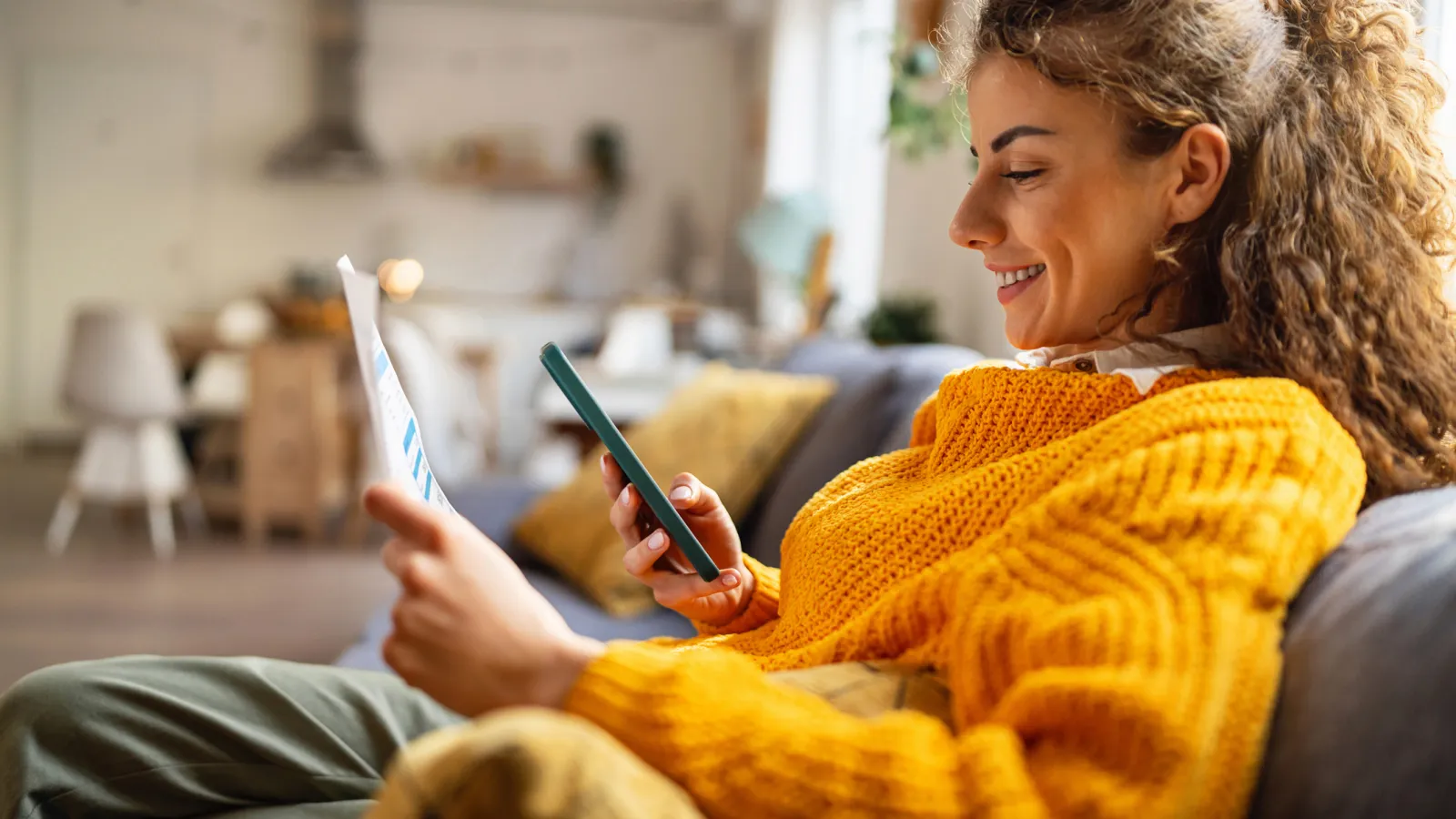Smiling woman in orange sweater lounging on couch using smartphone and holding a document in a cozy living room.