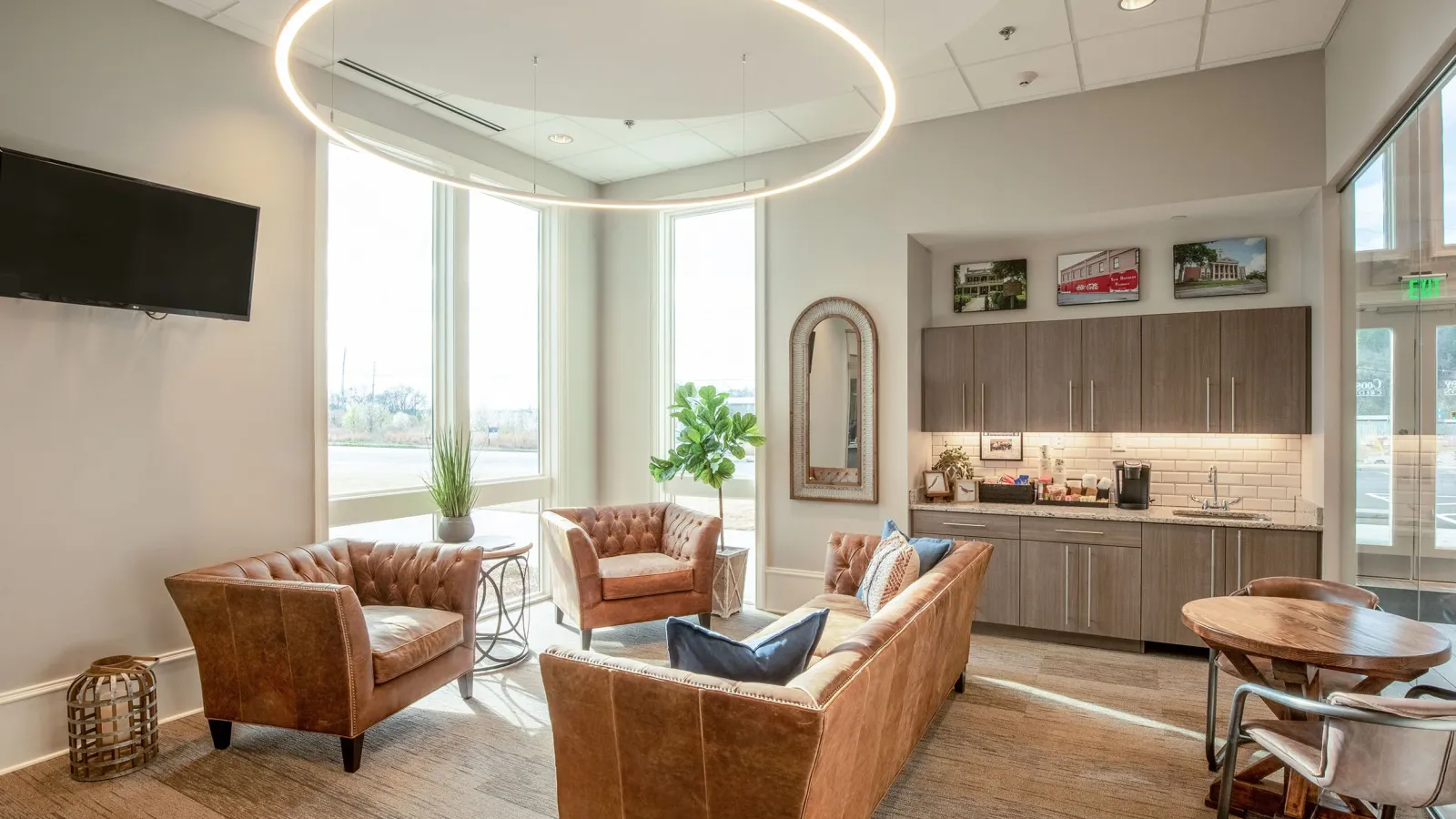 Bright modern lounge with leather sofas, circular LED ceiling light, kitchenette, and large windows providing natural light.