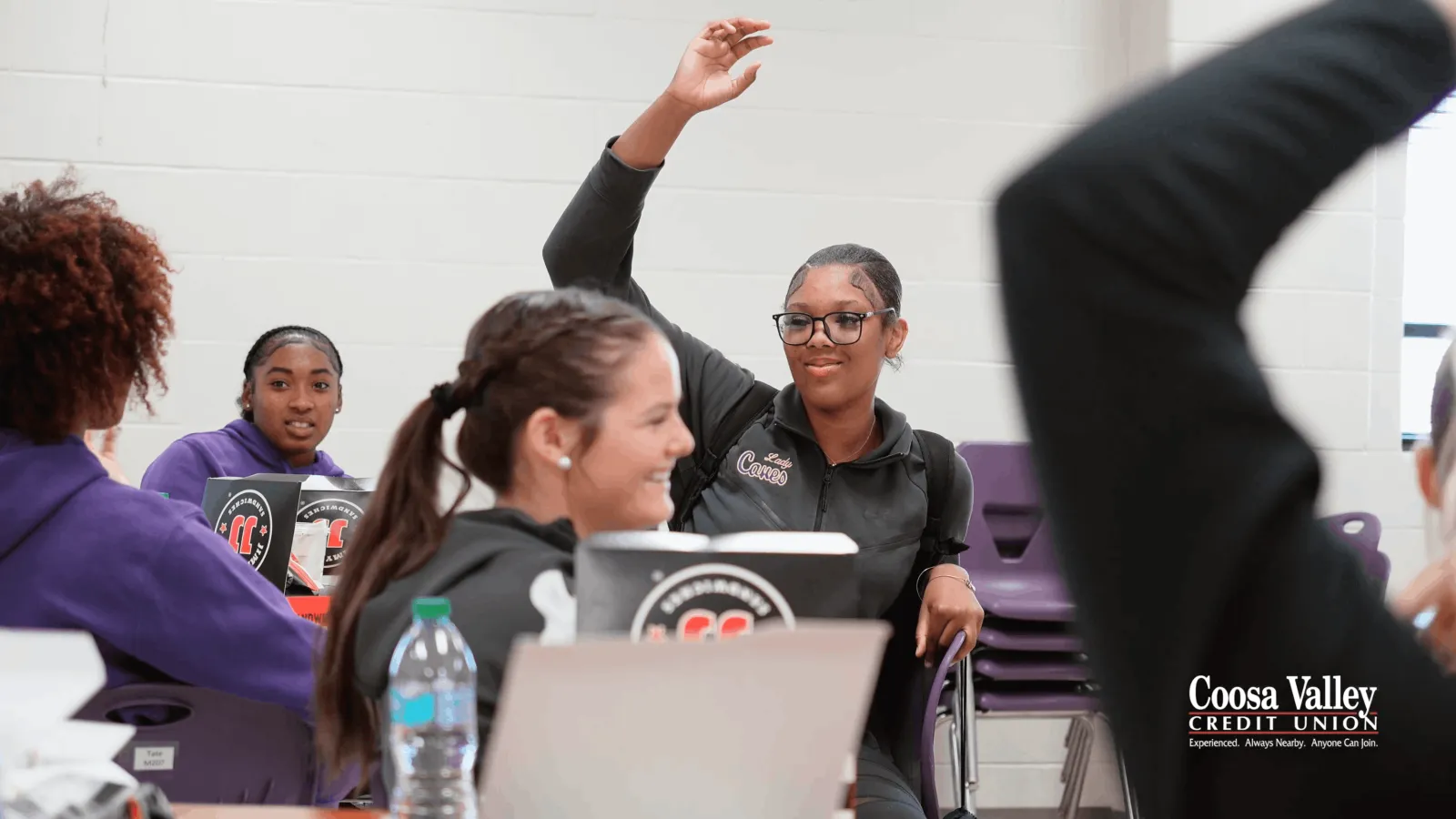 Students engaged in classroom discussion with one student raising hand and others smiling and listening attentively.