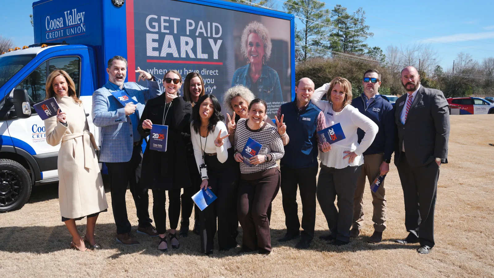 Group of smiling people posing outdoors in front of Coosa Valley Credit Union van with a Get Paid Early advertisement.