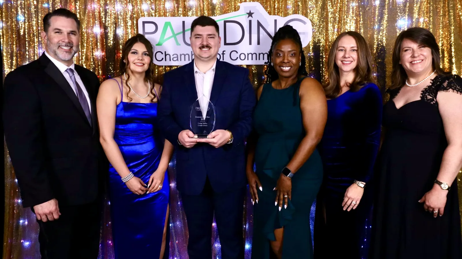 Group of six people dressed formally at an awards event with a gold sequin backdrop and Paulding Chamber of Commerce sign.