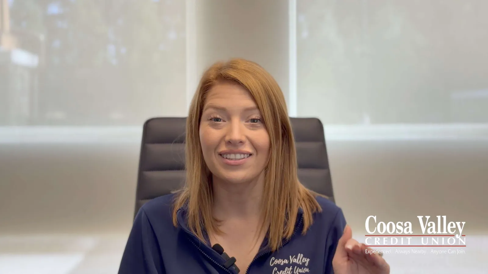 Smiling woman in blue Coosa Valley Credit Union jacket sitting in office chair with logo on bottom right.