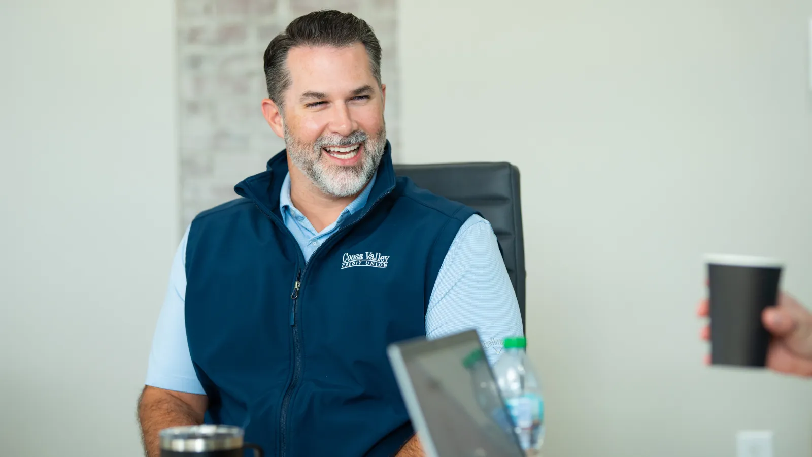 Smiling man in a blue vest sitting at a table during a casual meeting with a laptop and coffee cups.
