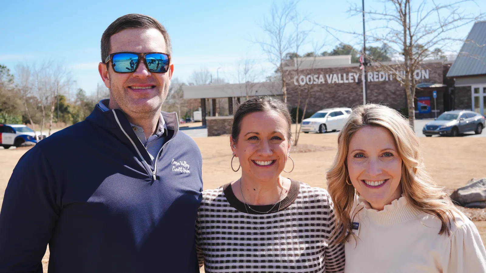 Three smiling people standing outdoors in front of Coosa Valley Credit Union building on a sunny day