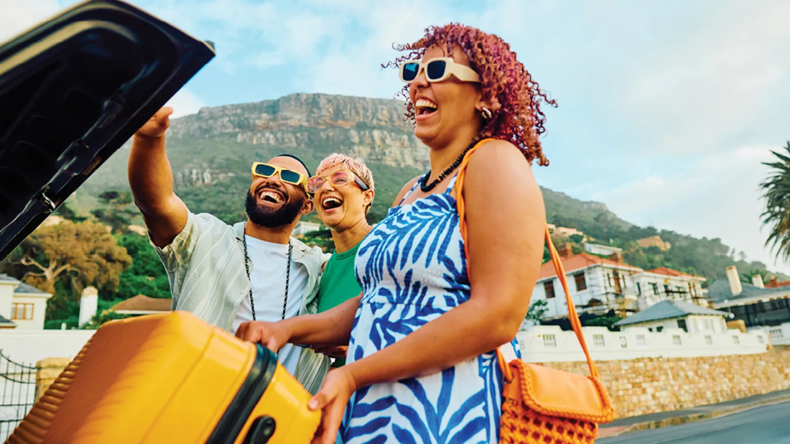 Three happy friends packing a yellow suitcase into a car trunk with mountain and houses in background
