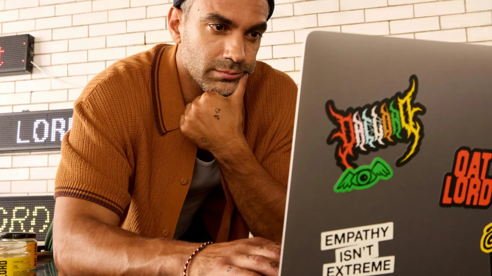Man wearing a beanie and brown shirt focused on laptop with stickers including 'Empathy Isn't Extreme'