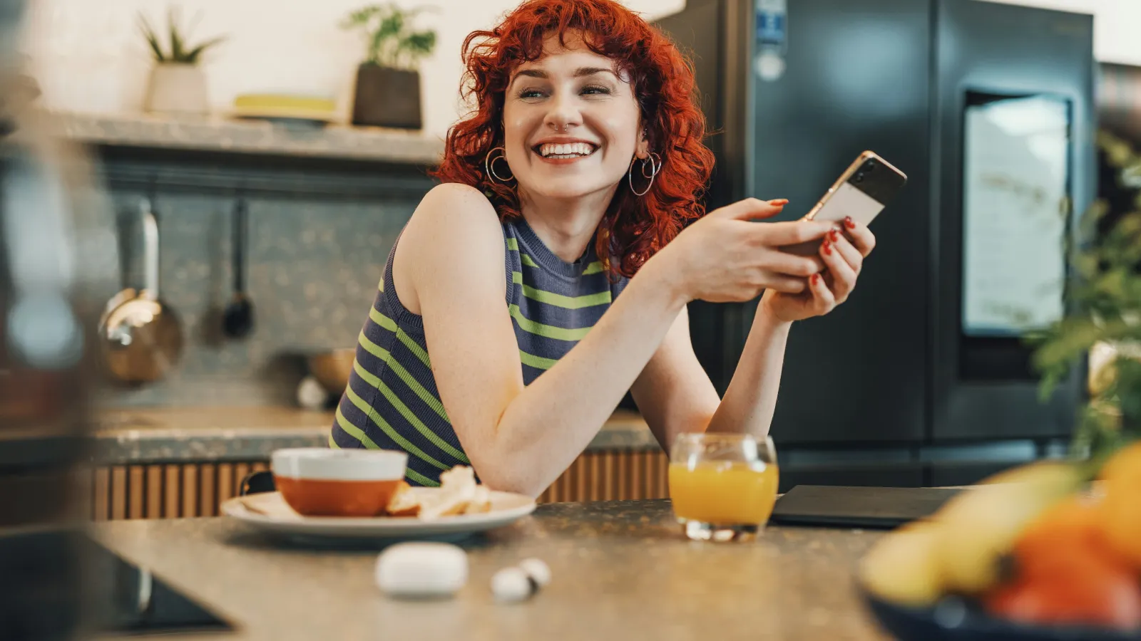 Smiling red-haired woman in striped tank top holding smartphone at kitchen counter with breakfast and juice.
