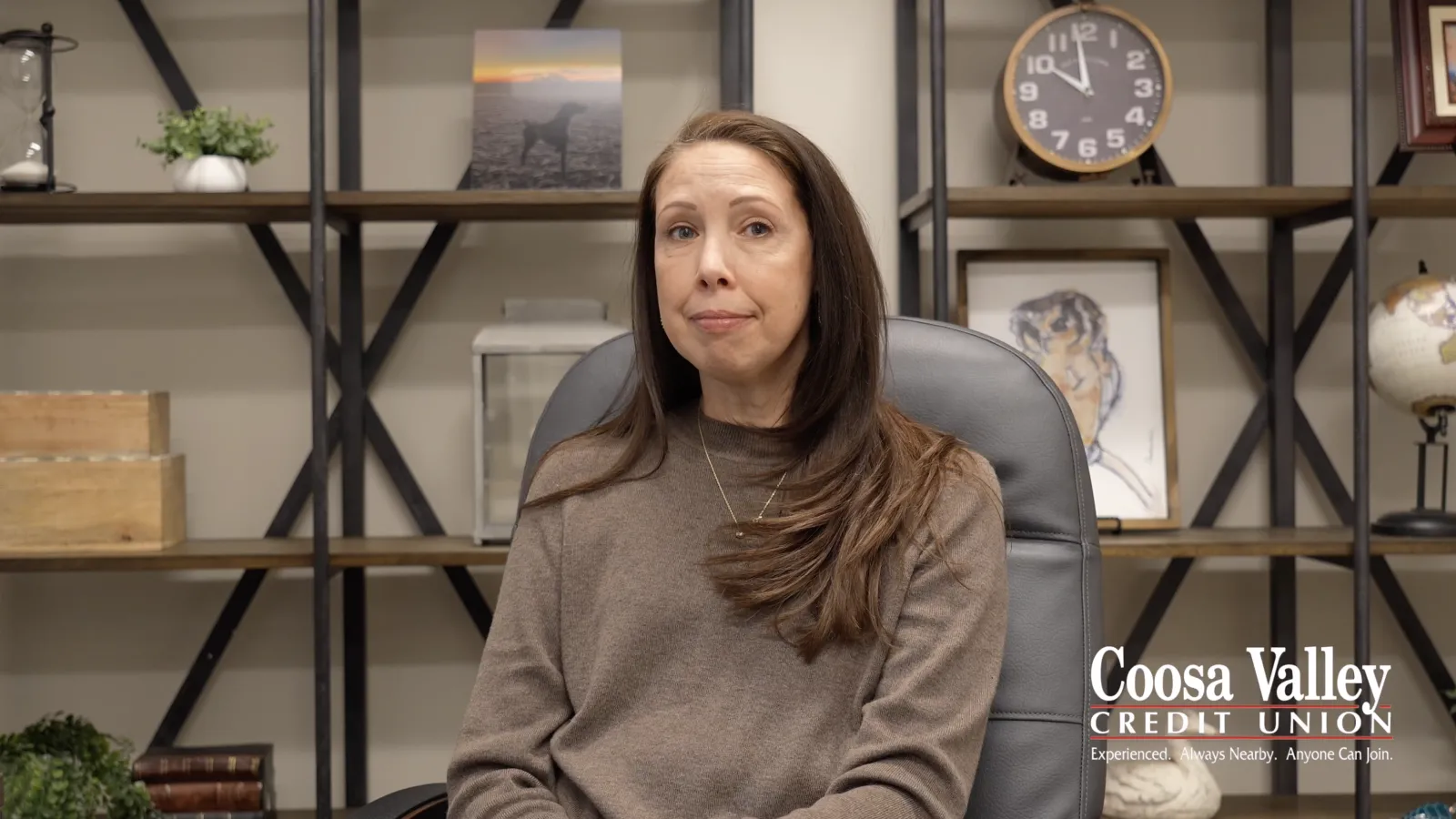 Woman with long brown hair sitting in an office chair in front of shelves with books and decorations.