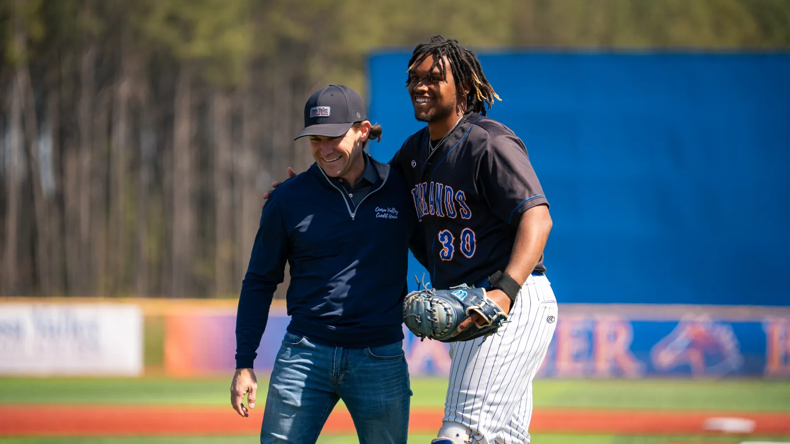 Two men smiling and embracing on a sunny baseball field with one in uniform and the other in casual wear.