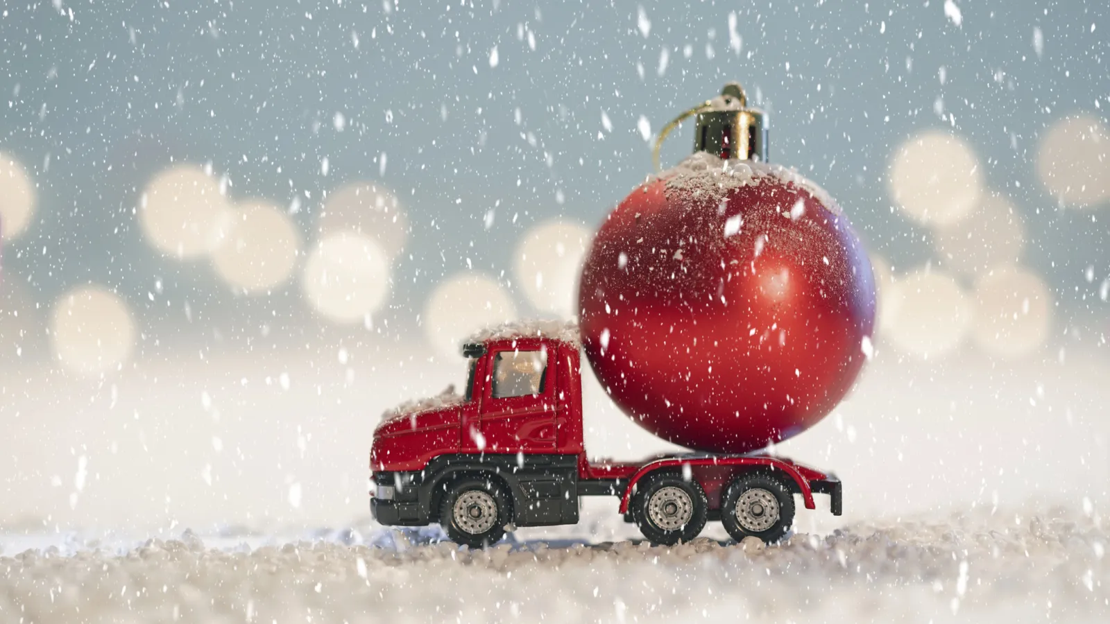 Red toy truck carrying a large red Christmas ornament in falling snow with blurred festive lights in background