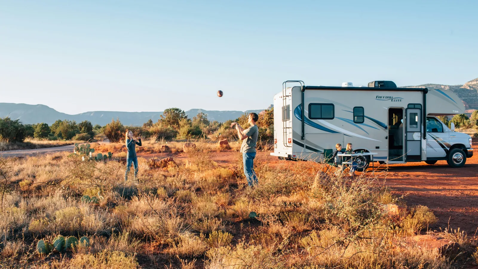 Family playing catch beside a parked RV in a scenic desert landscape with clear blue sky and mountains.