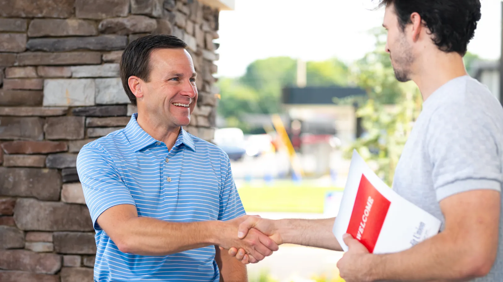 Two men shaking hands outside a building, one holding a welcome brochure, smiling and greeting.