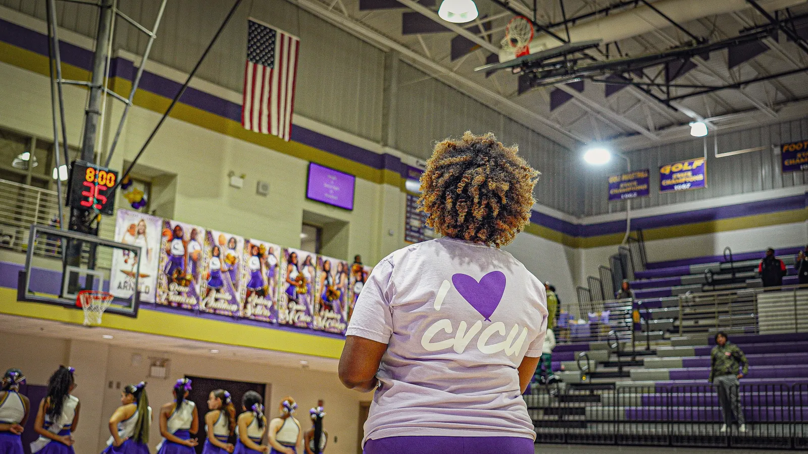 Woman wearing I love CVCU shirt watching cheerleaders in purple gymnasium with basketball hoop and American flag.