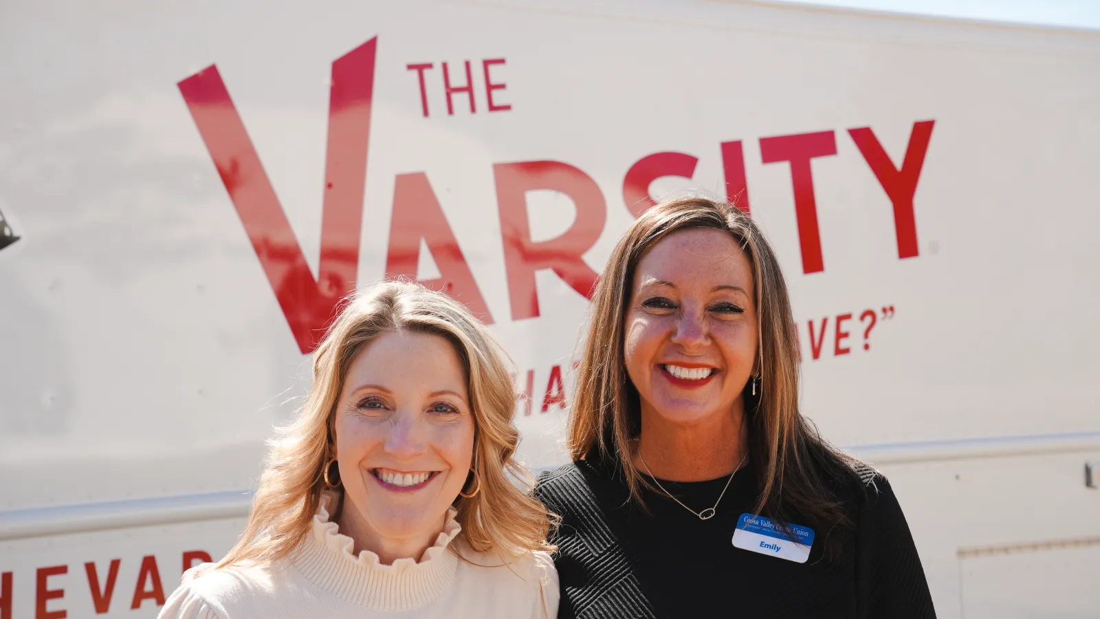 Two smiling women with name tags standing in front of The Varsity food truck on a sunny day