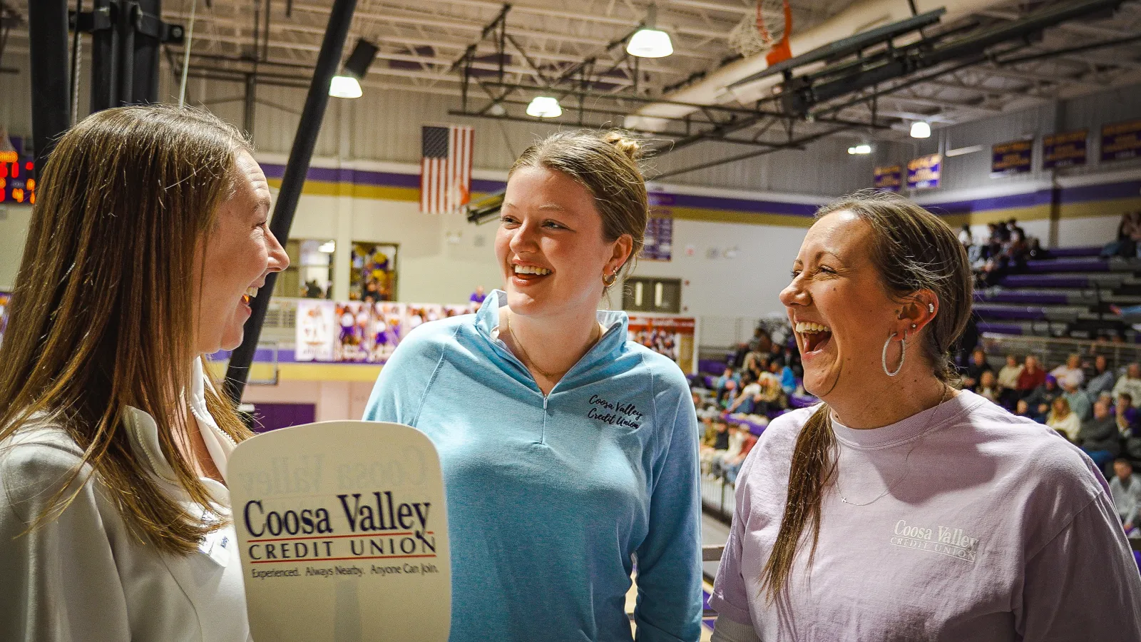 Three women smiling and talking indoors in a sports gym with Coosa Valley Credit Union signage visible.