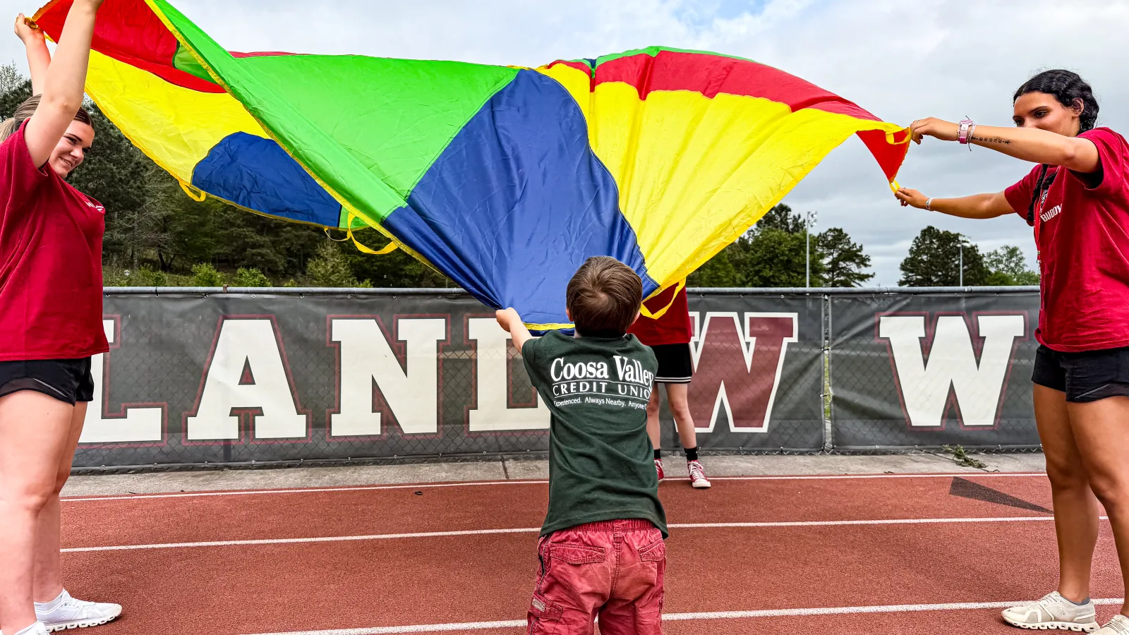 Children and adults playing with a colorful parachute on a running track in an outdoor sports area.