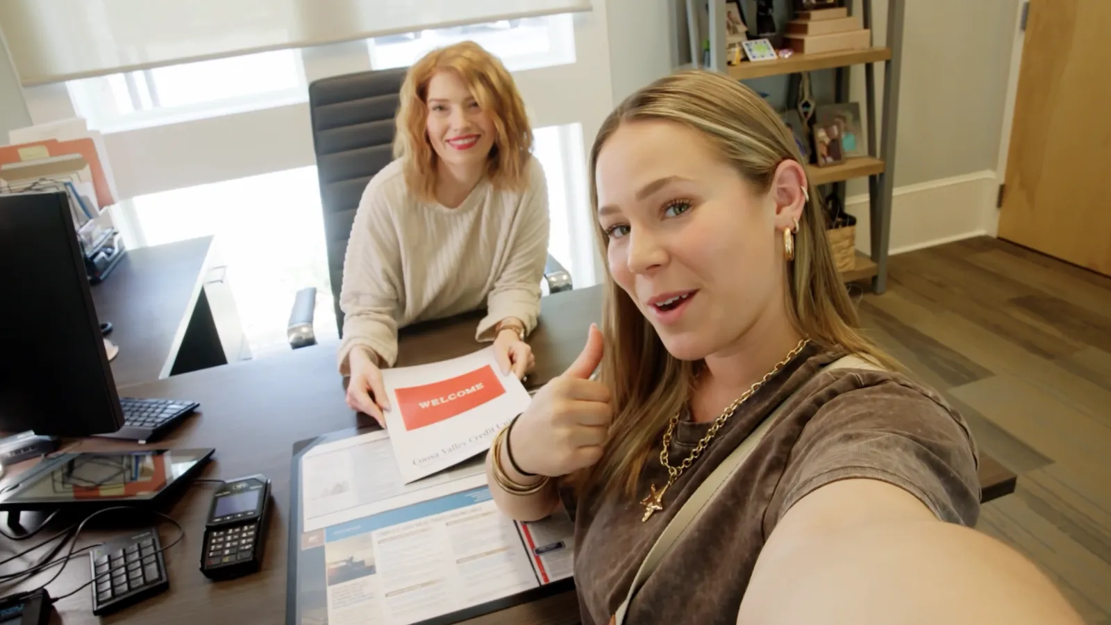 Two women in an office, one giving a thumbs up and the other holding a welcome brochure, smiling at the camera.