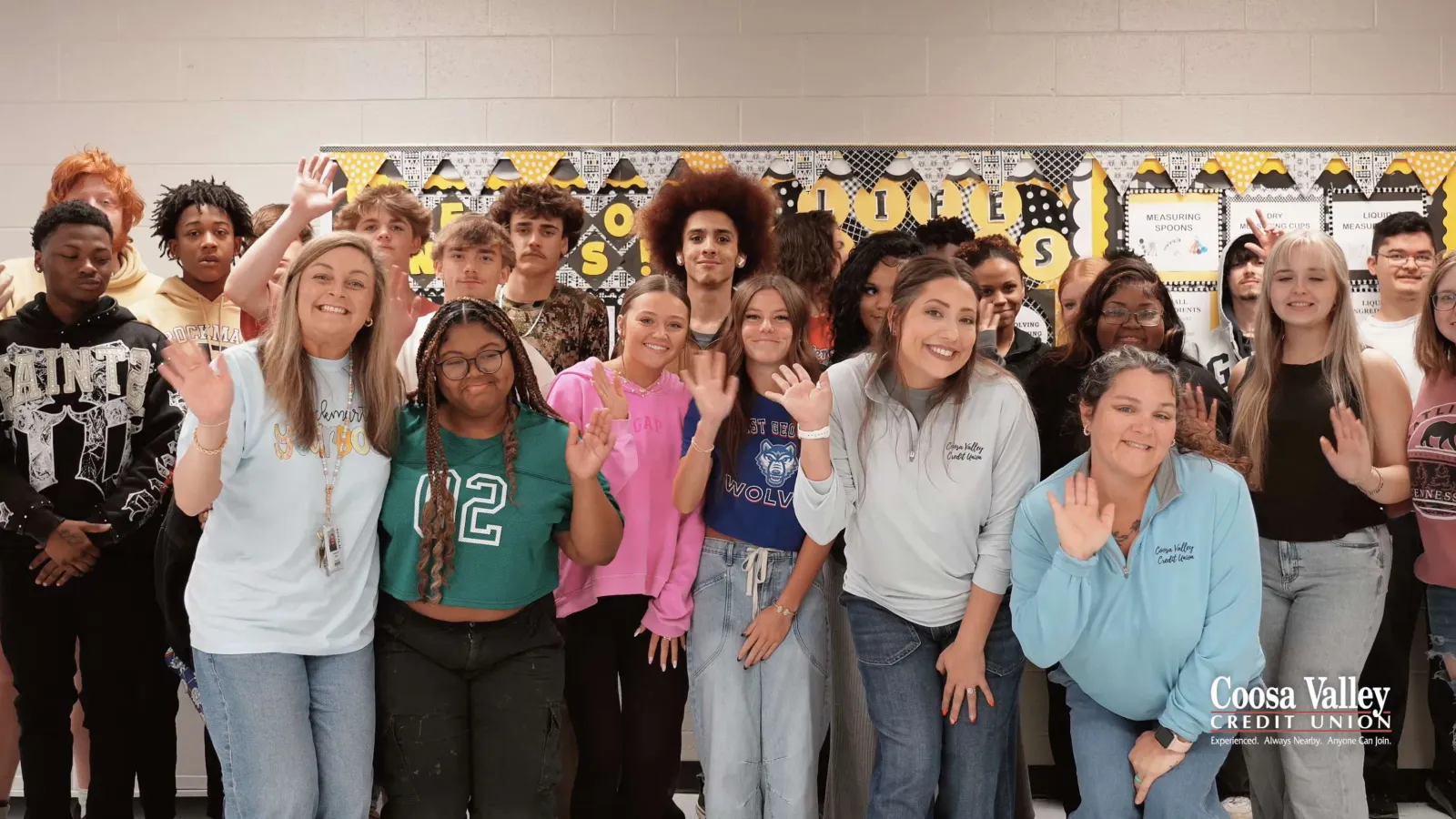 Diverse group of smiling students and adults waving in a classroom with bulletin board background and Coosa Valley logo.