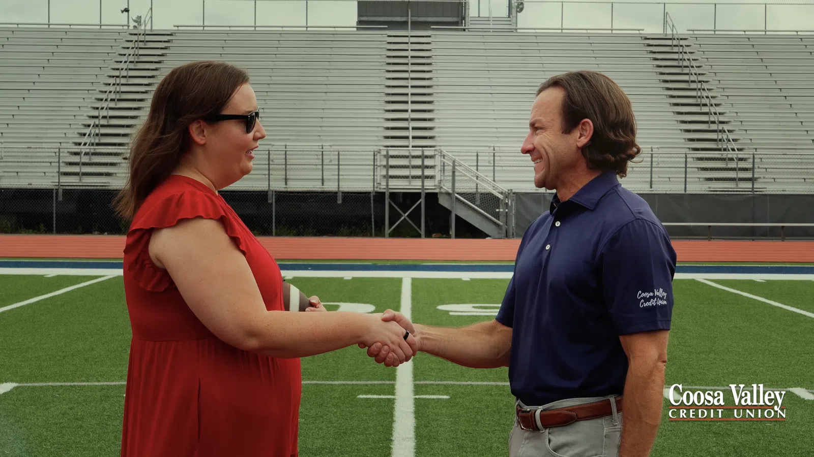 Two people shaking hands on a football field's center with empty bleachers and cloudy sky in background.