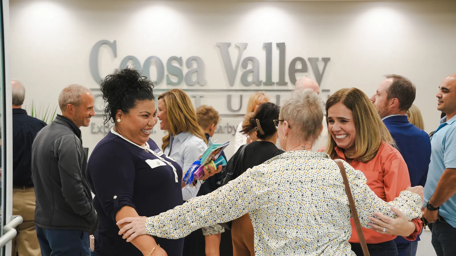 Smiling diverse group warmly greeting and embracing at Coosa Valley Community event in indoor setting.