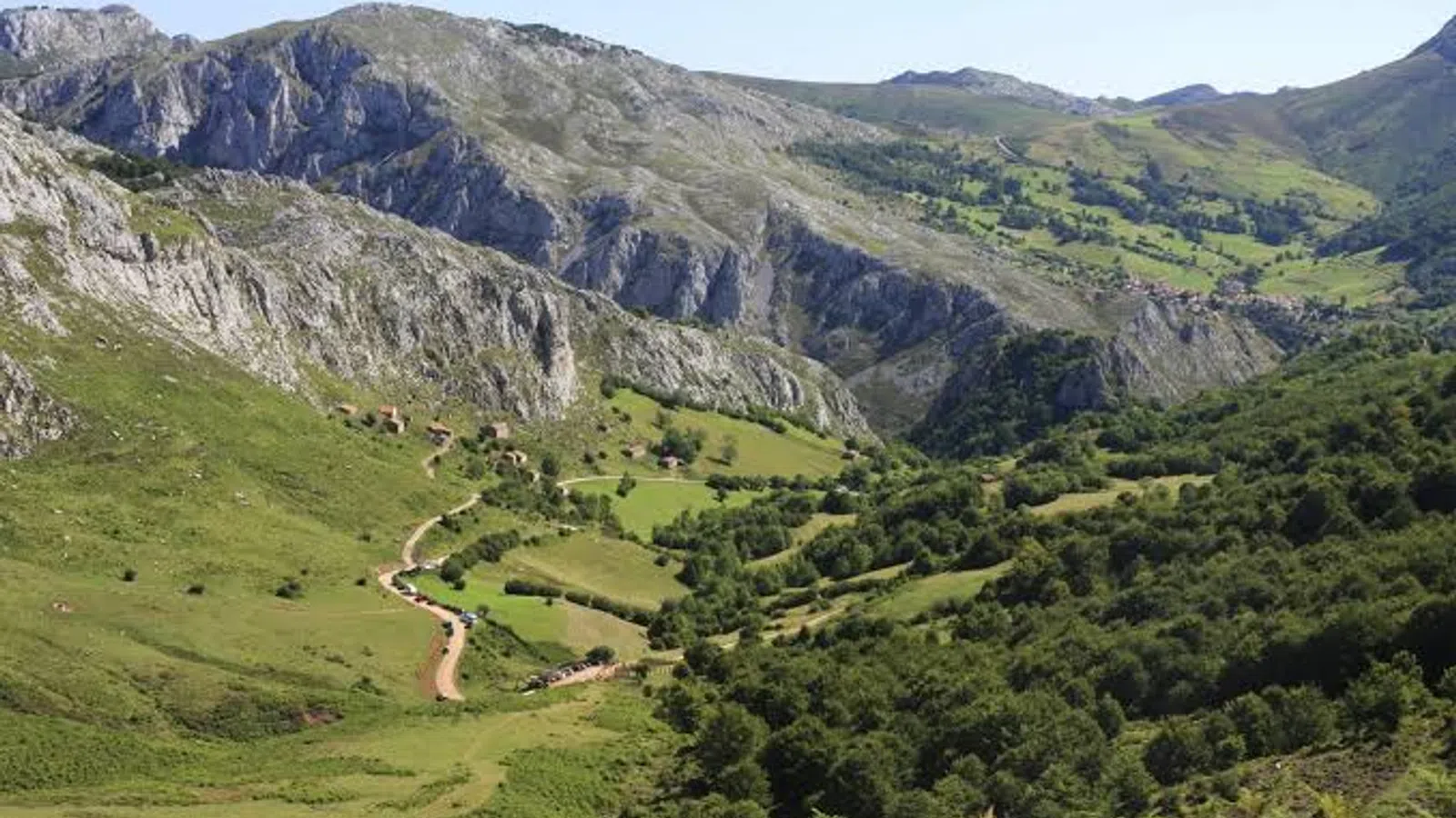 Mountain valley with green fields, rocky cliffs, trees, and a winding dirt path under clear blue sky