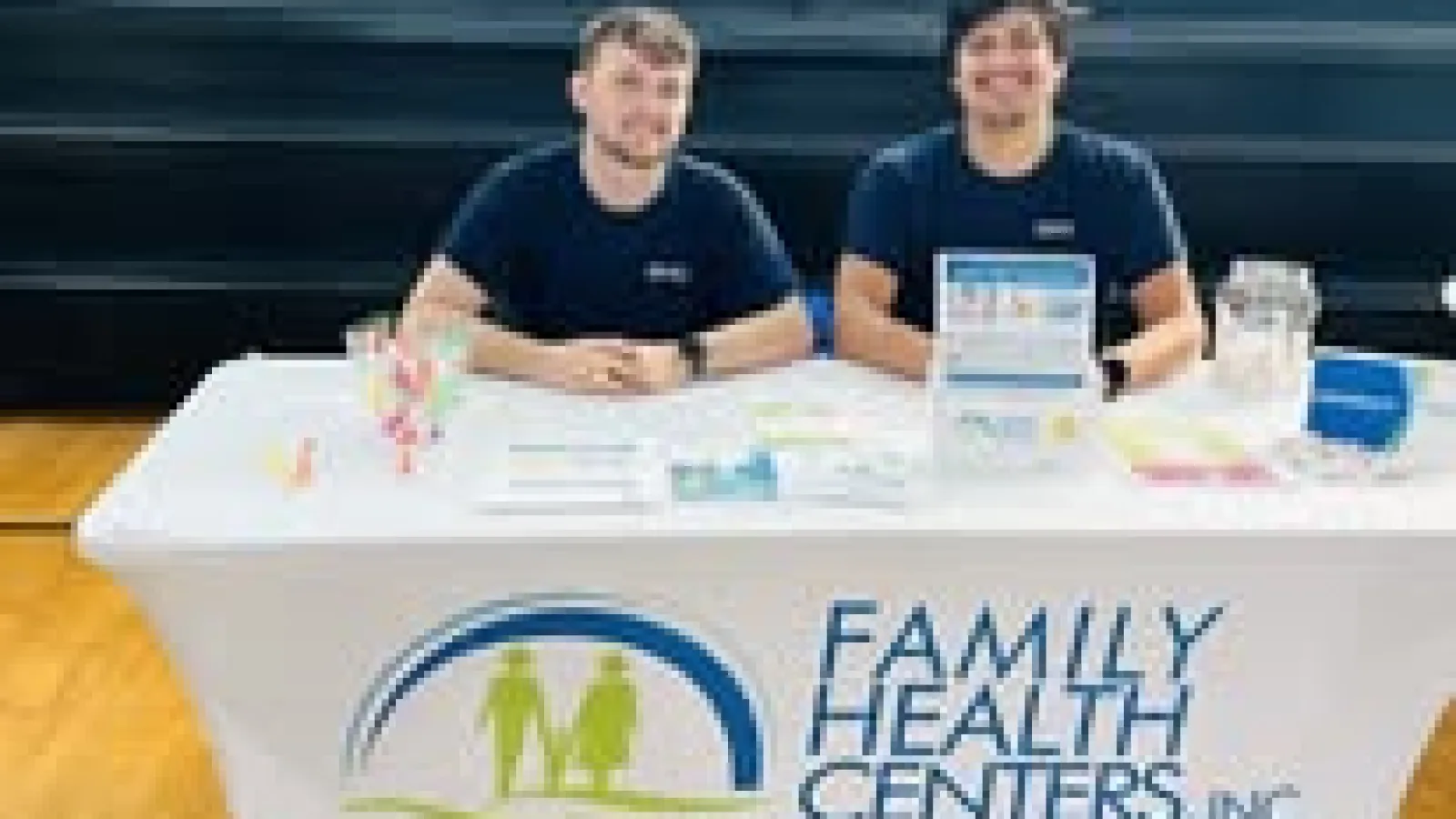 Two men sitting at a Family Health Centers promotional table with brochures and a donation jar indoors.