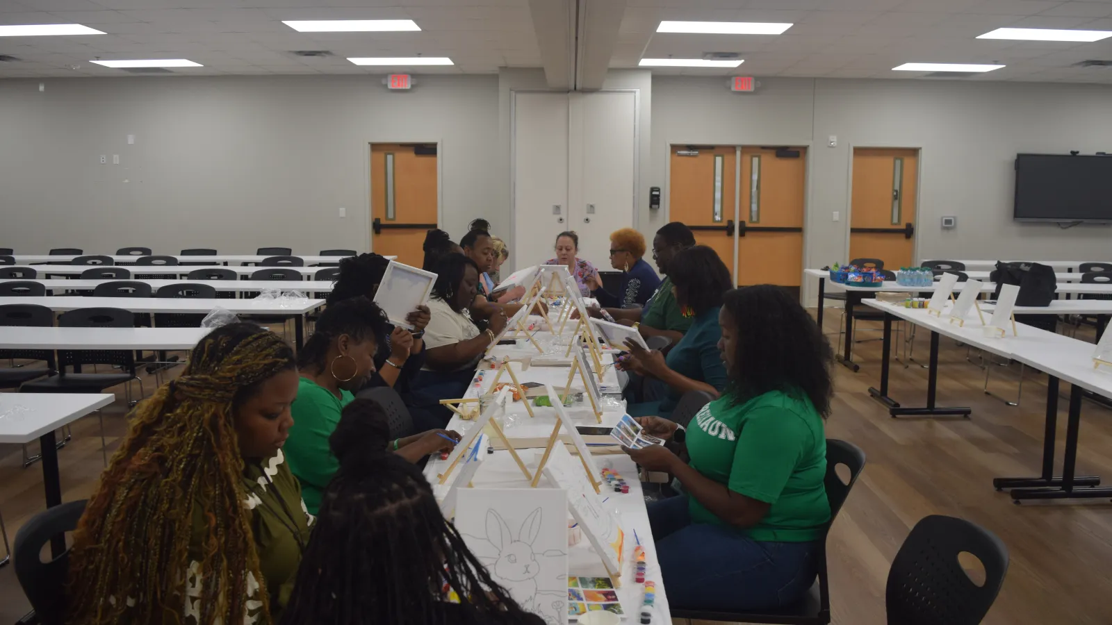 Group of women seated at a long table enjoying a painting class with easels and art supplies in a spacious room.