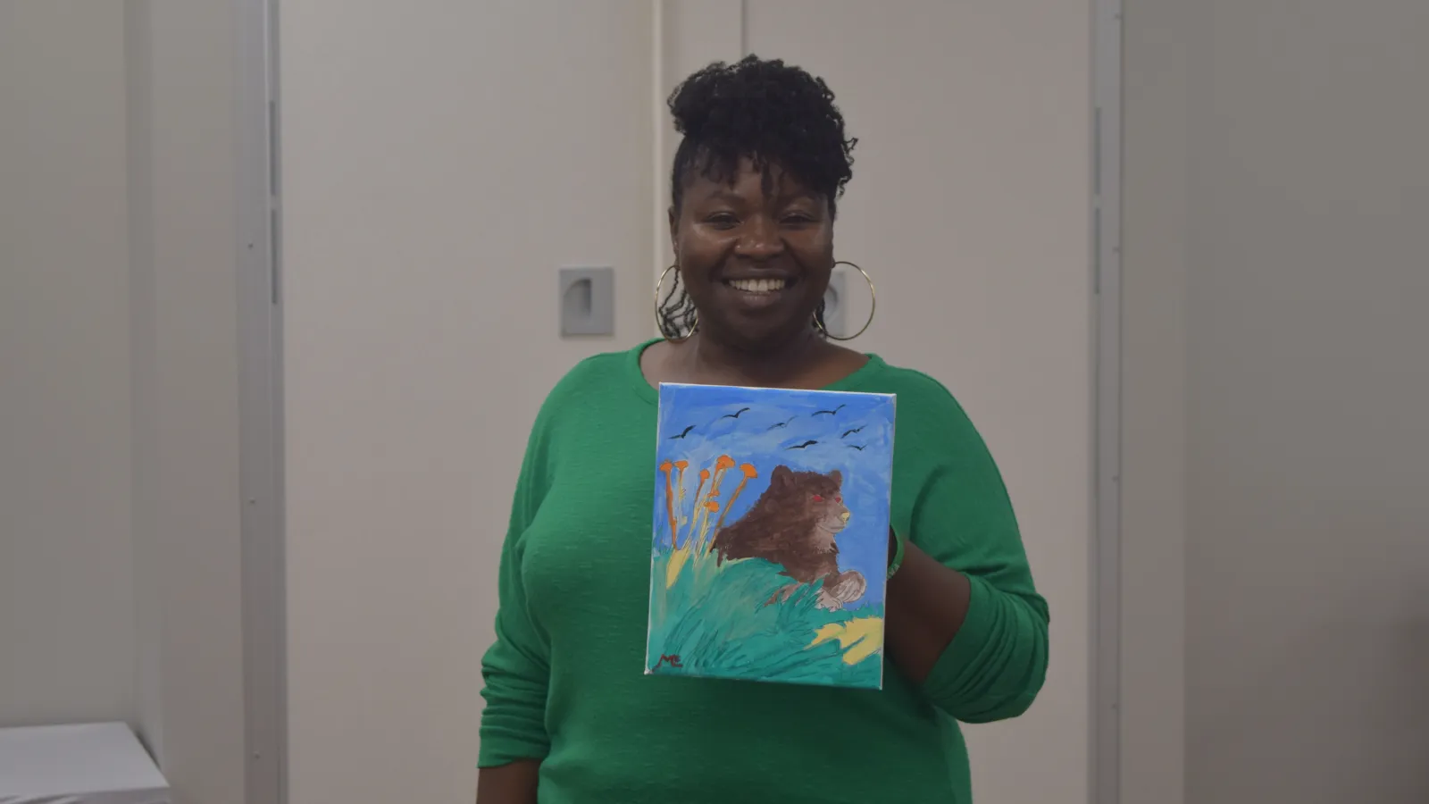 Smiling woman in green holding a colorful painting of a bear in nature with blue sky and birds.