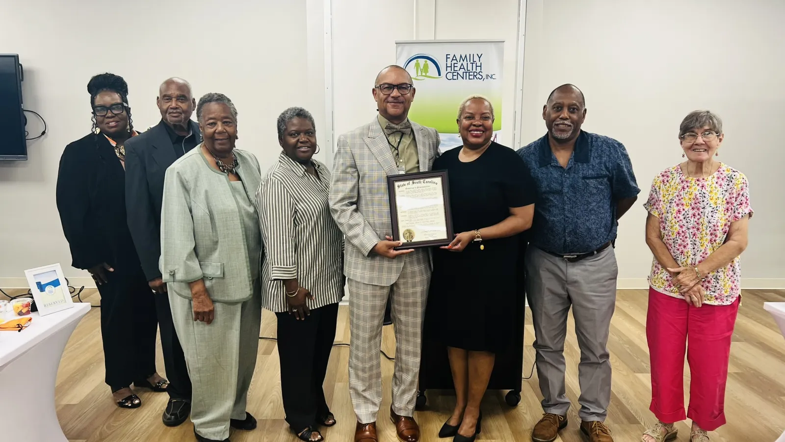 Group of diverse adults standing indoors with one man holding a framed certificate in front of a Family Health Centers banner.