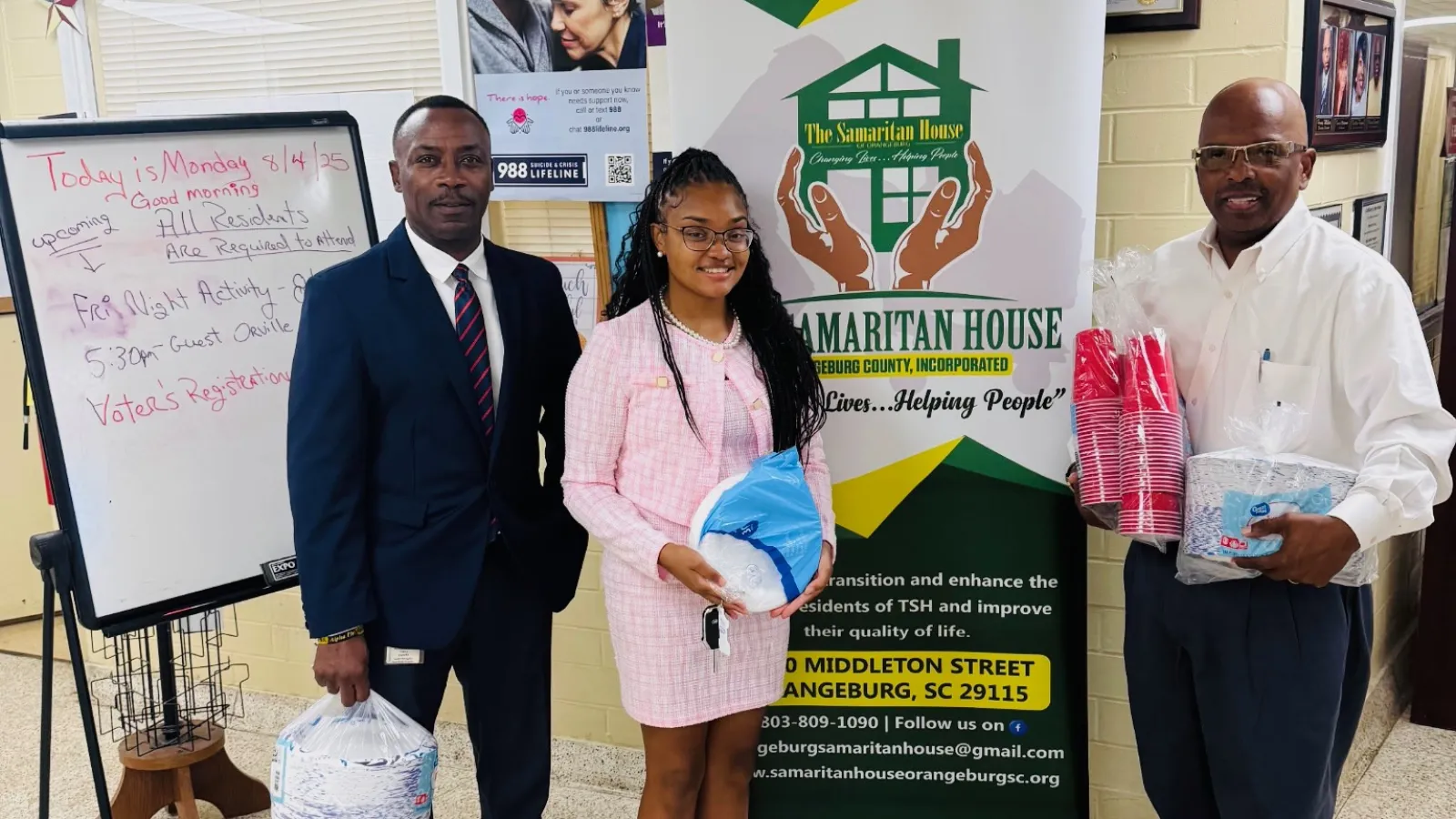 Three people standing indoors holding supplies in front of a Samaritan House banner for community support.