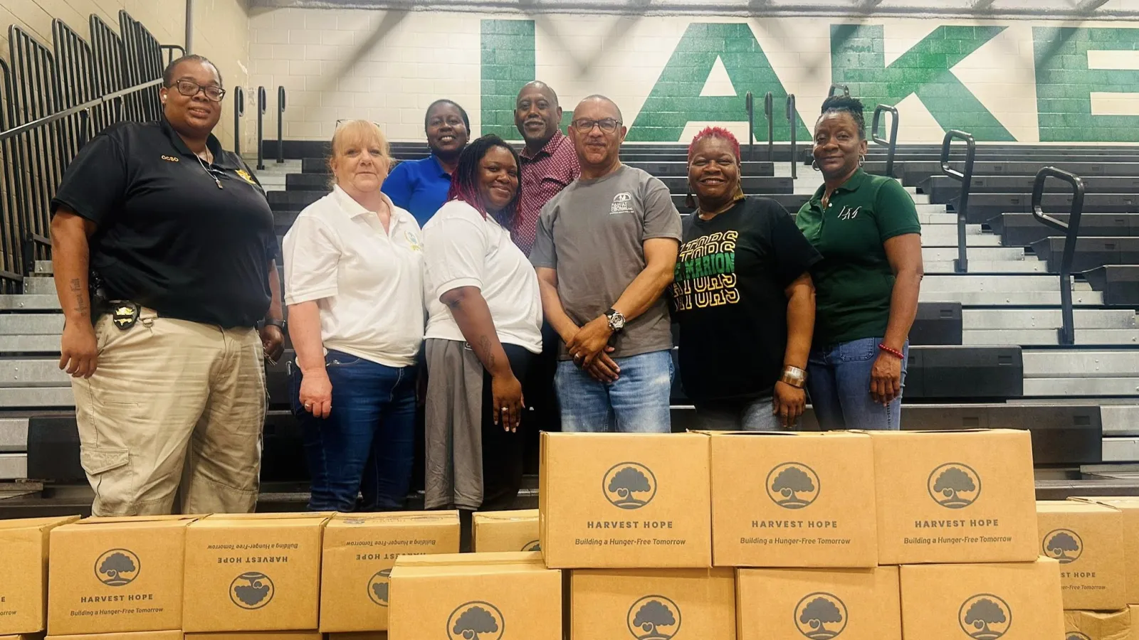 Group of eight people standing behind stacked Harvest Hope donation boxes in a gymnasium with bleachers and large green letters.
