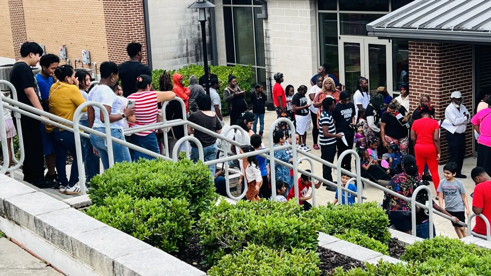 Diverse group of people waiting in a line outside a building with greenery and metal railings