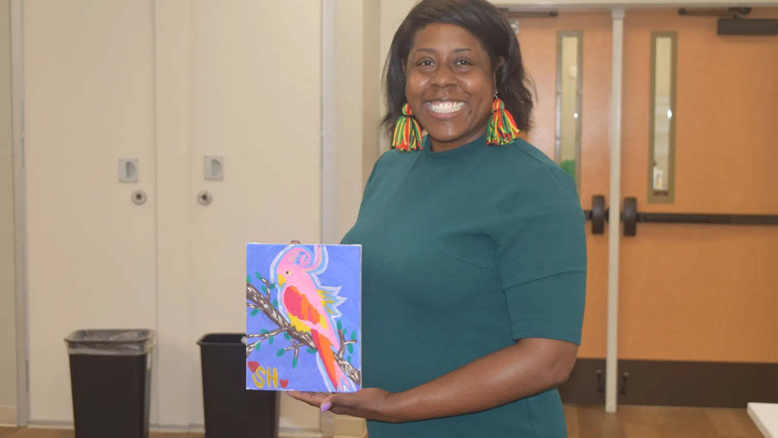 Smiling woman in green dress holding colorful parrot painting indoors with wooden floor and doors