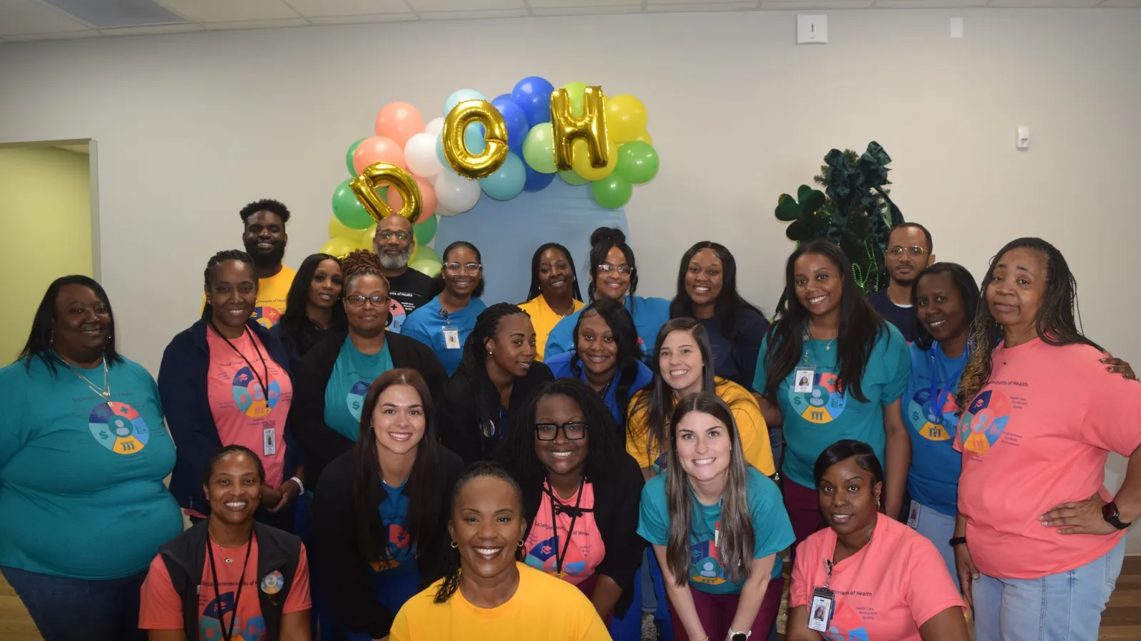 Group photo of smiling adults wearing colorful event t-shirts under balloons spelling DOH in a bright room.