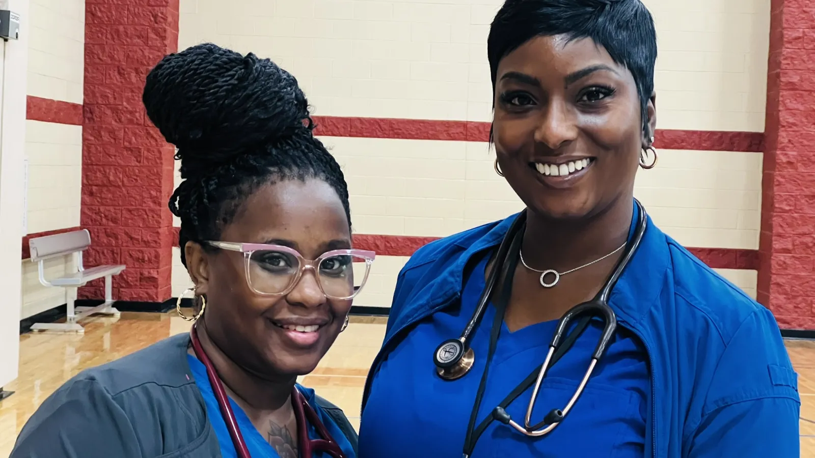 Two smiling female healthcare professionals wearing scrubs and stethoscopes in a gymnasium setting.